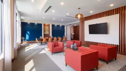 A spacious conference room with bold red and blue seating, a wall-mounted TV, and floor-to-ceiling windows letting in ample natural light.