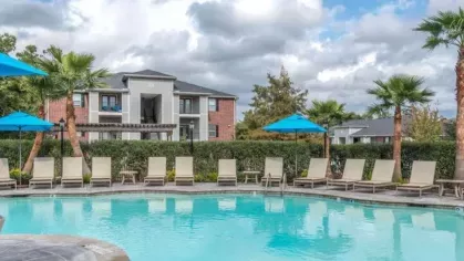 Resort-style swimming pool at The Landings of Conroe Luxury Apartments, surrounded by lush greenery, palm trees, and ample lounge seating under vibrant blue umbrellas.