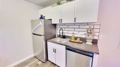Modern kitchen with sleek white cabinets, a subway tile backsplash, black countertops, and stainless steel appliances, offering a clean and functional design, seamlessly connected to a bright living space.