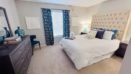 Elegant bedroom with a tufted headboard, cozy bedding, and soft carpeting, highlighted by decorative accents and ample natural light from a large window.