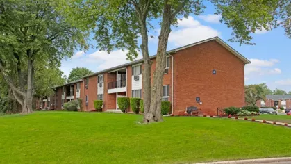 A red-brick apartment building with a large tree and a neatly landscaped lawn.