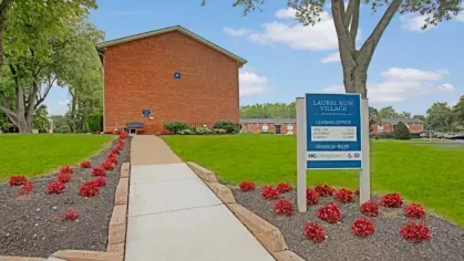 The walkway to the Laurel Run Village leasing office with a blue sign and well-manicured landscaping.