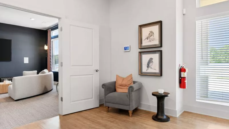A cozy seating area at Lawndale Commons Palm Luxury Apartments, featuring a gray armchair, side table, and framed bird artwork on a light-colored wall.