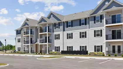 A modern apartment building at Lawndale Commons with elegant stone accents, white siding, and black balcony railings, surrounded by open parking spaces.