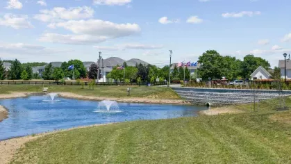 A scenic pond with two water fountains at Lawndale Commons Palm Luxury Apartments, surrounded by lush greenery, a stone bridge, and a well-manicured landscape.
