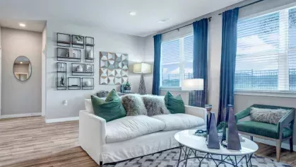 A stylish living room with a white couch, plush pillows, and geometric décor, complemented by large windows and a modern ceiling fan.