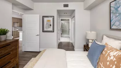 Spacious bedroom featuring a luxurious green velvet bedspread, elegant geometric artwork, and an adjoining walk-in closet. Modern lighting and stylish nightstands complete the space.