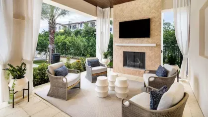 A luxurious covered seating area featuring a built-in fireplace, a mounted TV, and modern wicker chairs with blue cushions, surrounded by lush landscaping.