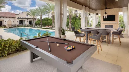 A covered outdoor lounge with a billiards table, bar seating, and a fireplace, overlooking a sparkling resort-style pool at Locklyn West Palm.