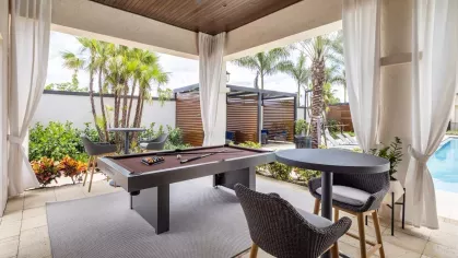 A luxurious cabana featuring a billiards table, high-top seating, and a tranquil view of the landscaped pool area.