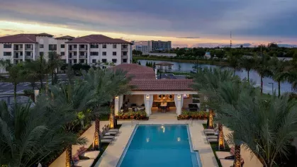 A sunset aerial view of Locklyn West Palm, showcasing the resort-style pool, poolside lounge, and Mediterranean-style apartment buildings with a scenic lake backdrop.