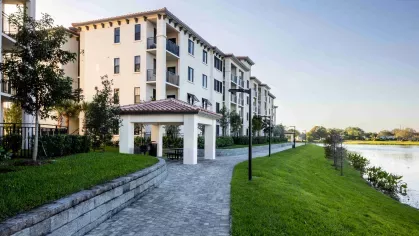 A paved walkway lined with lush greenery runs alongside the modern, Mediterranean-inspired apartment buildings, offering residents a peaceful lakeside retreat.