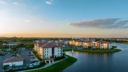 A panoramic view of Locklyn West Palm, highlighting waterfront apartment buildings with terracotta roofs, surrounded by greenery and tranquil water features under a clear sky.