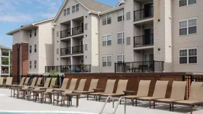 Sunlit swimming pool area featuring tan lounge chairs and contemporary apartment buildings with private balconies in the background.