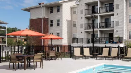Sunlit swimming pool area featuring tan lounge chairs and contemporary apartment buildings with private balconies in the background.