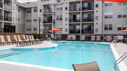 Sunlit swimming pool area featuring tan lounge chairs and contemporary apartment buildings with private balconies in the background.