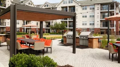A modern pergola-covered dining space with built-in stainless steel grills and a long table with orange-accented chairs.