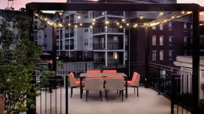 A covered dining area with string lights and vibrant orange chairs, offering a cozy atmosphere with scenic apartment views at dusk.