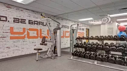 A modern fitness center featuring a weight rack with a variety of dumbbells, a strength training machine, and a motivational mural with bold text on a white brick wall.
