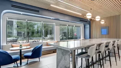 A stylish communal workspace with a marble bar-height table, plush blue lounge chairs, and floor-to-ceiling windows.