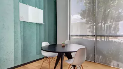 A cozy meeting room with a round black table, white chairs, a glass whiteboard, and a teal accent wall beside a large sunlit window.