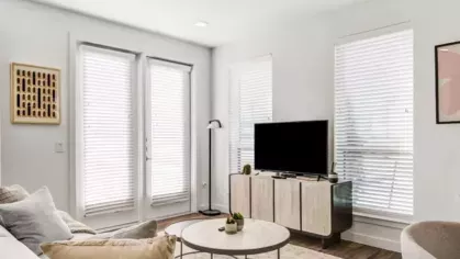 A bright living space with a beige couch, a round coffee table, and large windows with white blinds.