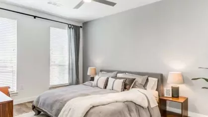 A spacious bedroom with a modern gray upholstered bed, neutral bedding, and warm lighting from two bedside lamps.