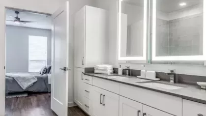 A bright bathroom with a double vanity, illuminated mirrors, and sleek white cabinetry leading to a bedroom.