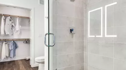A modern glass-enclosed shower with a rain showerhead, adjacent to a walk-in closet with hanging clothes.