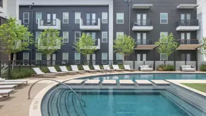 A modern apartment pool area with lounge chairs, lush landscaping, and multi-story residential buildings in the background.