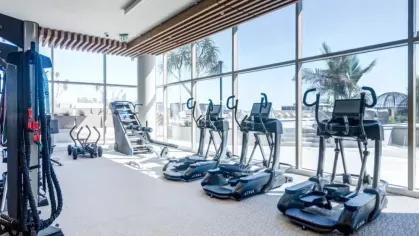 A bright fitness room with ellipticals and cardio machines overlooking a cityscape through floor-to-ceiling windows.