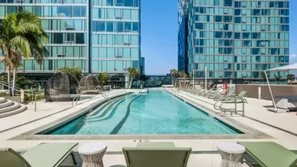 Modern outdoor pool area surrounded by sleek glass apartment buildings with green lounge chairs and palm trees.