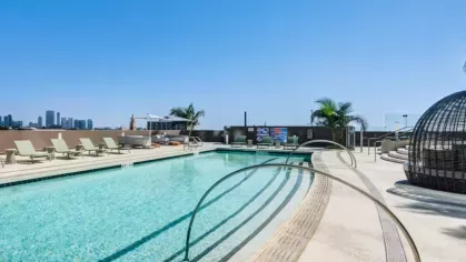 Rooftop pool area with a clear blue pool, green lounge chairs, and a view of the city skyline under a sunny sky.