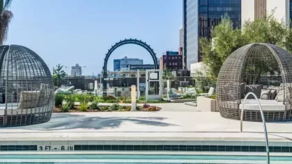Poolside seating with wicker egg-shaped cabanas, lush greenery, and city buildings in the background.