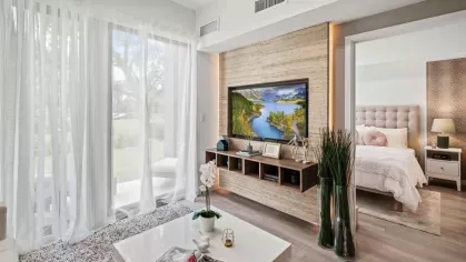A contemporary apartment living area featuring floor-to-ceiling windows with sheer curtains, a built-in entertainment center, and a cozy bedroom visible in the background.