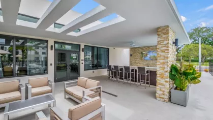 A stylish covered outdoor lounge at Oak Enclave Luxury Apartments, featuring cushioned chairs, a stone accent bar, and a pergola roof design for an inviting entertainment space.