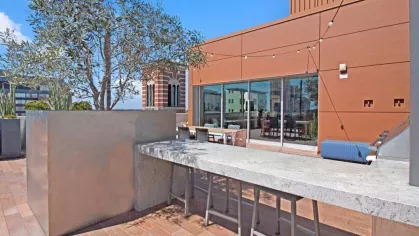 "Outdoor dining and entertainment space at The Pacific Luxury Apartments, showcasing a long concrete bar, natural greenery, and a striking orange accent wall."