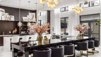 A luxurious communal space featuring a black dining table with velvet chairs, elegant gold chandeliers, and large windows with natural light.