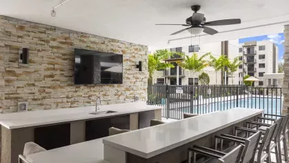 An outdoor bar and entertainment area with a stone accent wall, a flat-screen TV, and a view of the swimming pool at Pine Ridge Luxury Apartments.