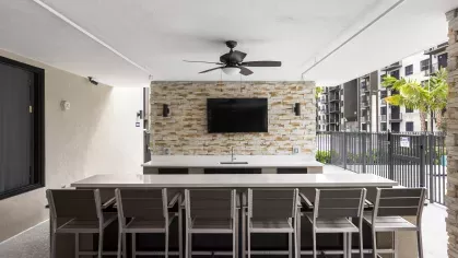 A communal outdoor space with a bar, seating, and a stone wall featuring a mounted flat-screen TV, offering a view of the surrounding apartment buildings.