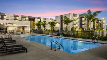 A pristine swimming pool in front of a modern clubhouse with lounge chairs arranged around the pool deck, showcasing luxurious amenities at Pine Ridge Luxury Apartments.