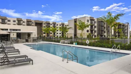 A pristine swimming pool in front of a modern clubhouse with lounge chairs arranged around the pool deck, showcasing luxurious amenities at Pine Ridge Luxury Apartments.