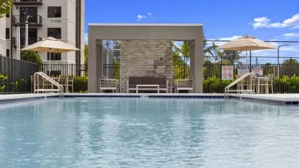 A pristine swimming pool in front of a modern clubhouse with lounge chairs arranged around the pool deck, showcasing luxurious amenities at Pine Ridge Luxury Apartments.
