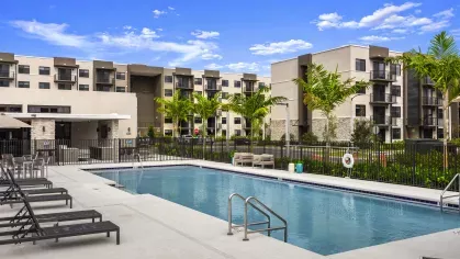 A pristine swimming pool in front of a modern clubhouse with lounge chairs arranged around the pool deck, showcasing luxurious amenities at Pine Ridge Luxury Apartments.