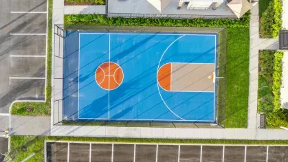 Aerial view of a vibrant blue and orange basketball court at Pine Ridge Luxury Apartments, surrounded by green landscaping and adjacent to parking spaces.