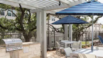 Outdoor grilling and dining area at Vista At Town Green, featuring a stainless steel grill, pergola-covered seating, and blue patio umbrellas for a comfortable outdoor experience.
