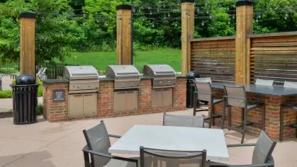 Outdoor grilling station at Riverworks Luxury Apartments, featuring multiple stainless steel grills, ample seating, and a natural green backdrop.