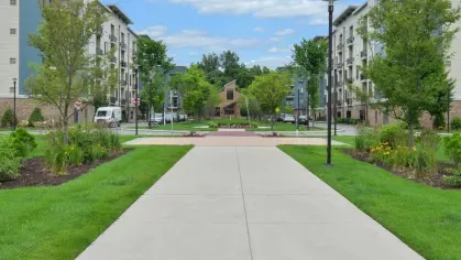 Wide concrete pathway leading through the manicured lawns and green spaces of Riverworks Luxury Apartments, surrounded by modern residential buildings and natural landscaping.