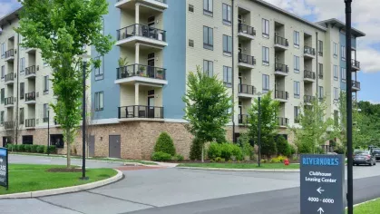 The exterior view of Riverworks Luxury Apartments showcases modern architecture with a mix of blue and cream siding, private balconies, and lush landscaped surroundings near the clubhouse leasing center.