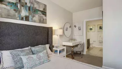 A well-appointed bedroom featuring a stylish upholstered headboard, contemporary artwork, and a writing desk with a decorative mirror, leading to an en-suite bathroom.